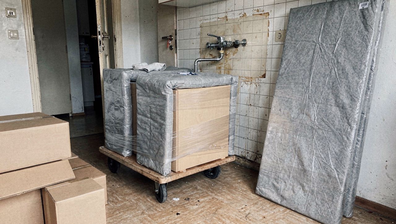 Abgebaute Küche in Umzugsdecken auf einem Rollbrett, dahinter die leere Wand mit Wasseranschluss